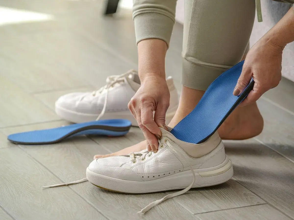 Manos insertando una plantilla azul en una bota blanca. Otra plantilla y una bota están en el suelo.