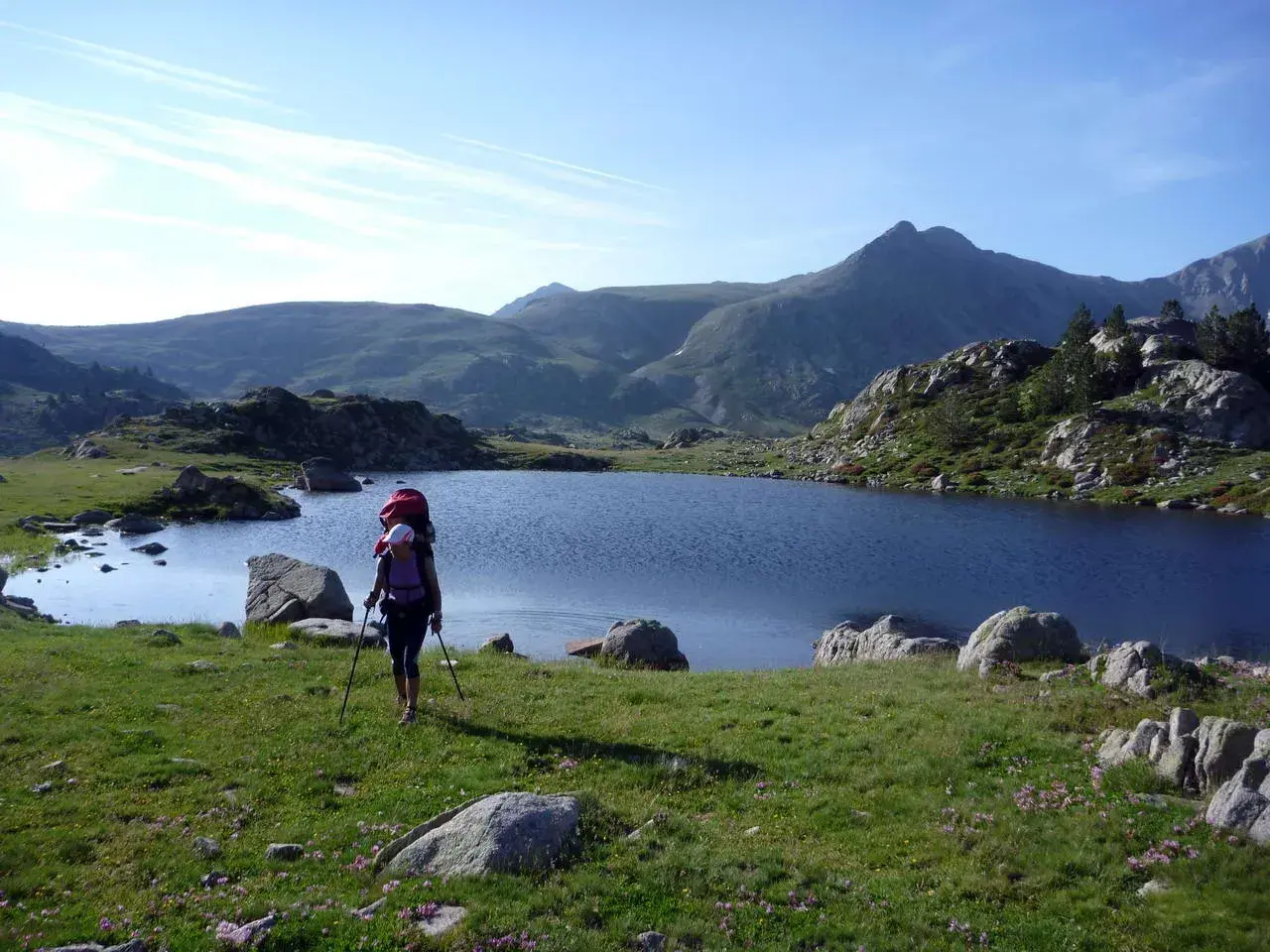 Senderista con mochila y bastones camina por un prado verde hacia un lago azul en los Pirineos de GRP Andorra.