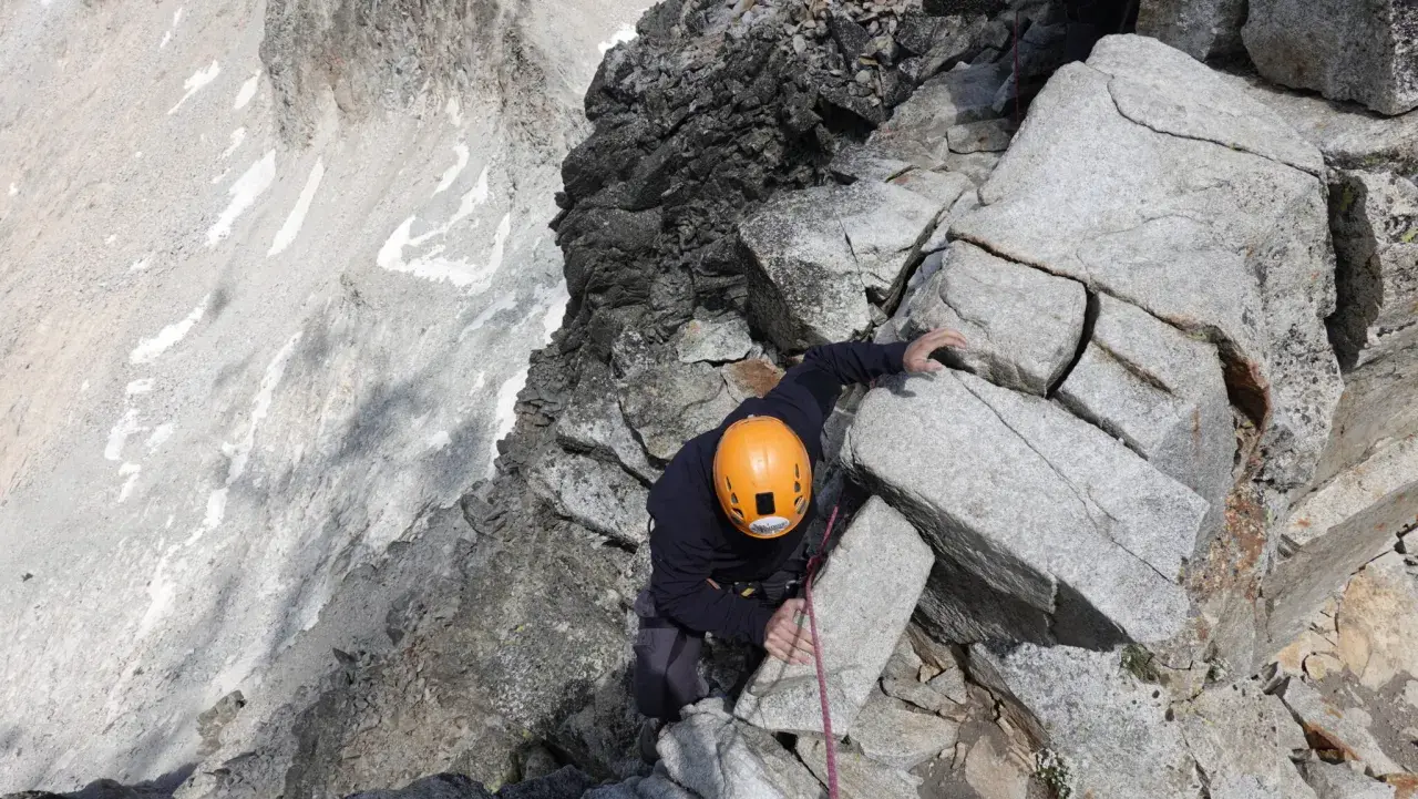 Alpinista con casco naranja escala roca, buscando el aneto por coronas.