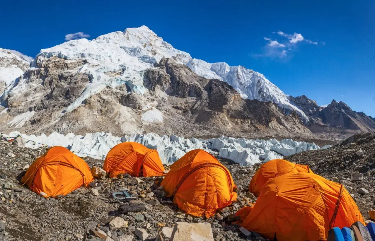 Campamento de expedición en la base de la cordillera más alta del mundo, con tiendas naranjas y glaciares imponentes bajo un cielo azul.