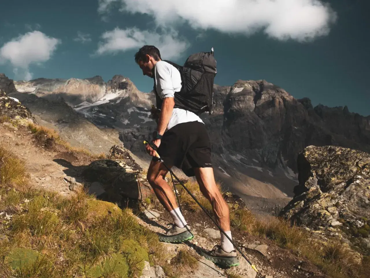 Kilian Jornet asciende una ladera rocosa en los Pirineos, con bastones y mochila, bajo un cielo azul con nubes.