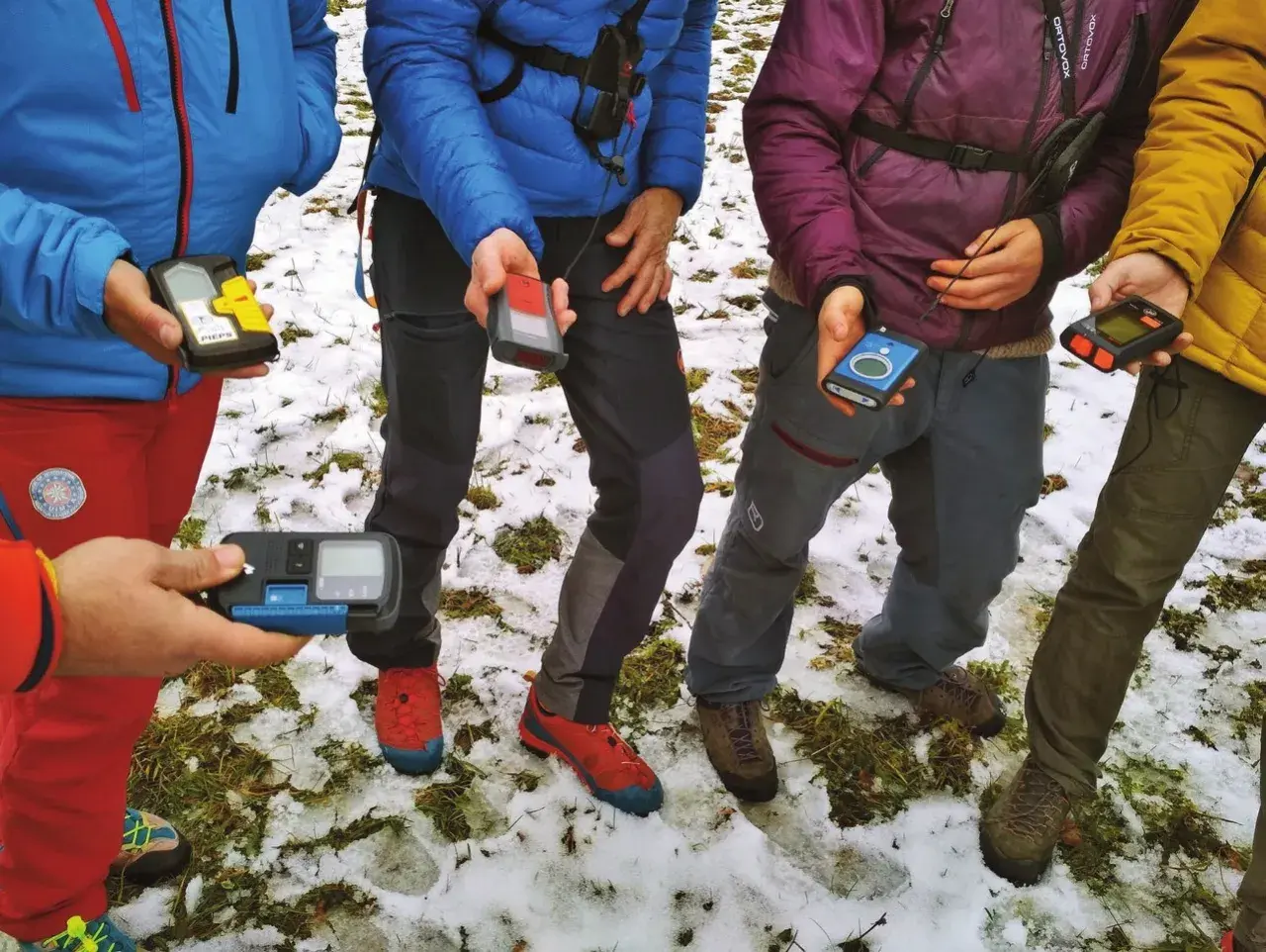 Personas en la nieve sostienen varios dispositivos de búsqueda de avalanchas, como el PIEPS, para una lección de seguridad en montaña.