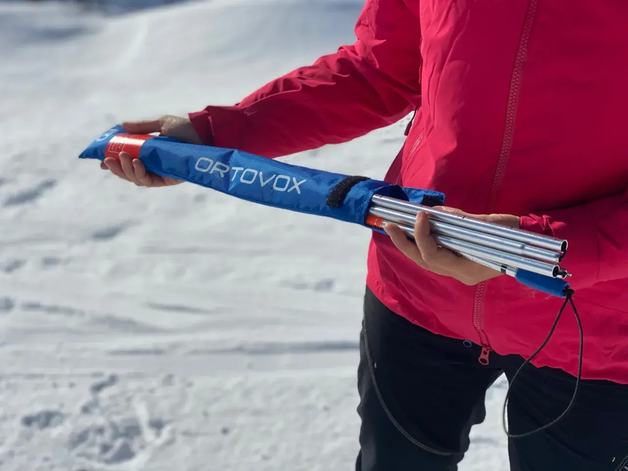 Persona con chaqueta rosa sostiene una arva, sonda y pala ORTOVOX en una bolsa azul, lista para la aventura en la nieve.