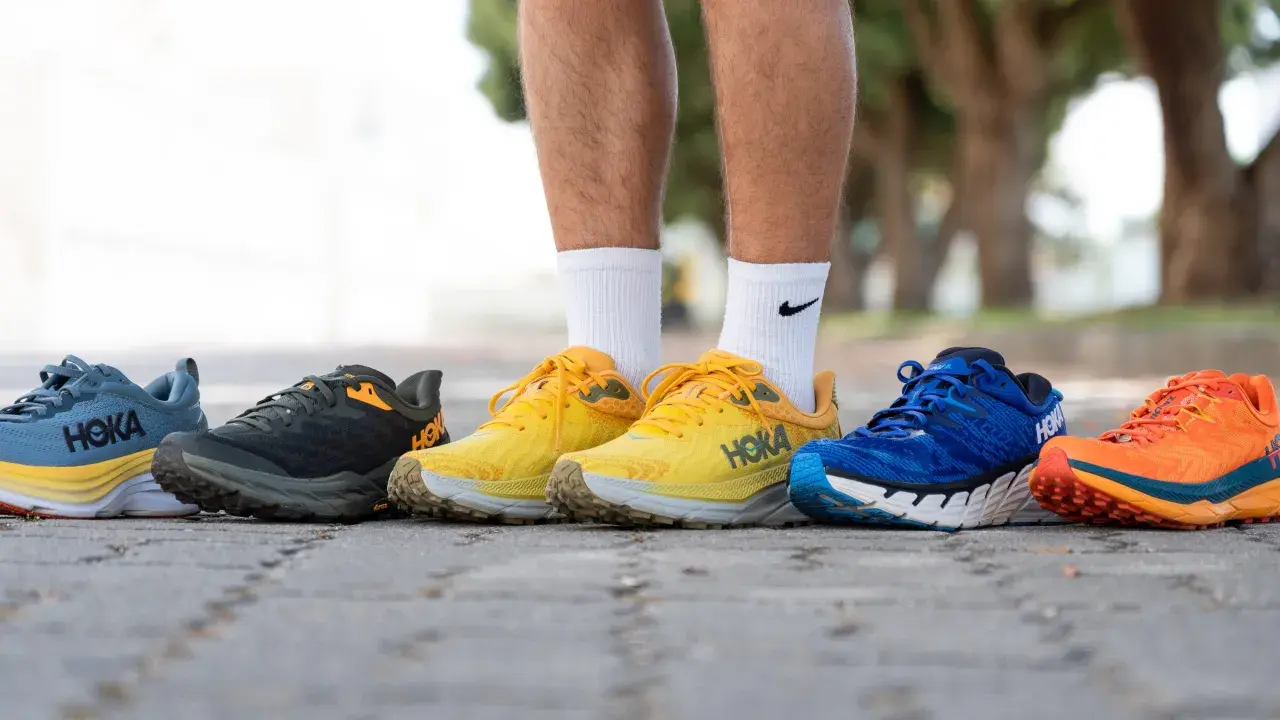 Cinco pares de zapatillas Hoka con gran amortiguación, en colores azul, gris, amarillo, azul y naranja, alineados en un camino.
