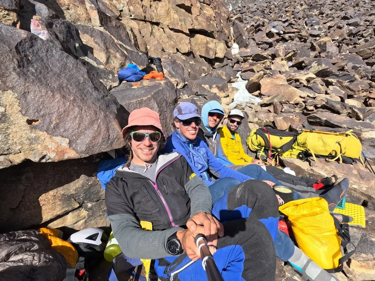 Cuatro montañistas sonríen en la ladera rocosa, listos para la aventura. El **bru busom** de uno de ellos, un sombrero rosa, destaca entre el equipo de expedición.