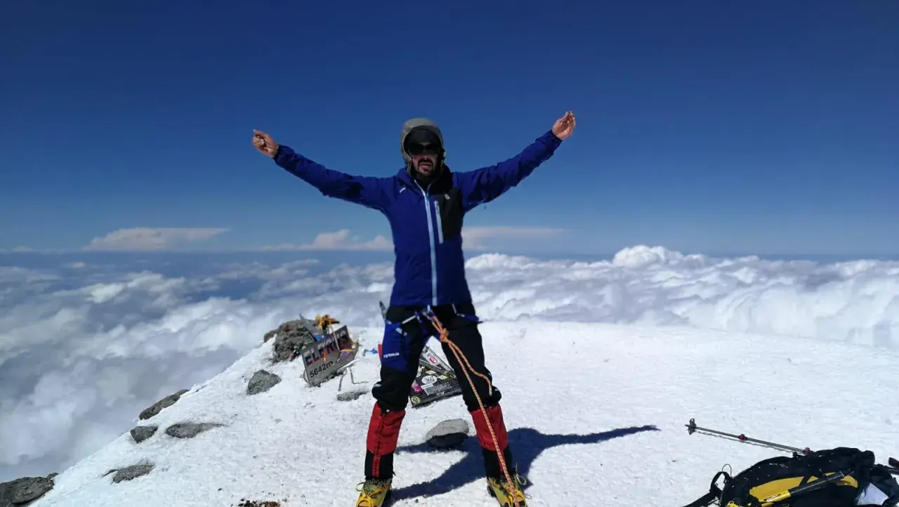Javier García Díaz, alpinista, celebra en la cima nevada, rodeado de nubes y un cielo azul intenso.