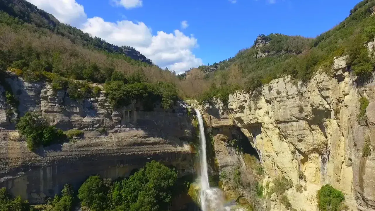 Cascada impresionante en un cañón rocoso, ideal para senderismo en Cataluña. El agua cae en una cortina blanca sobre la piedra dorada.