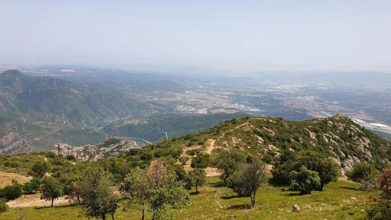 Vista panorámica desde Sant Miquel de Montserrat. Montañas verdes y un pueblo se extienden hasta el horizonte brumoso.