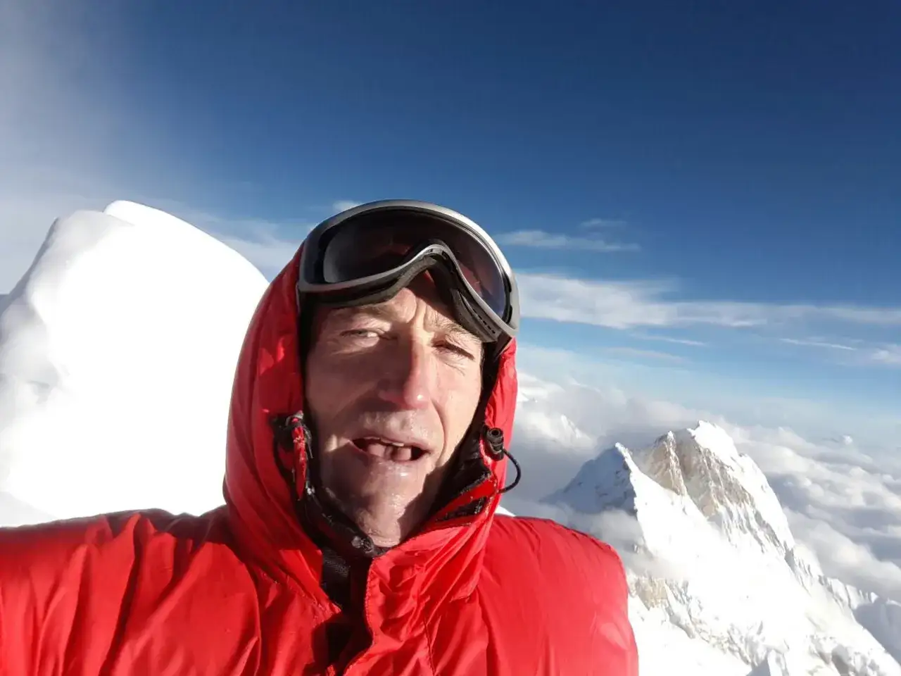 Alberto Zerain, con gafas de esquí y chaqueta roja, sonríe en la cima de una montaña nevada, rodeado de nubes y un cielo azul intenso.