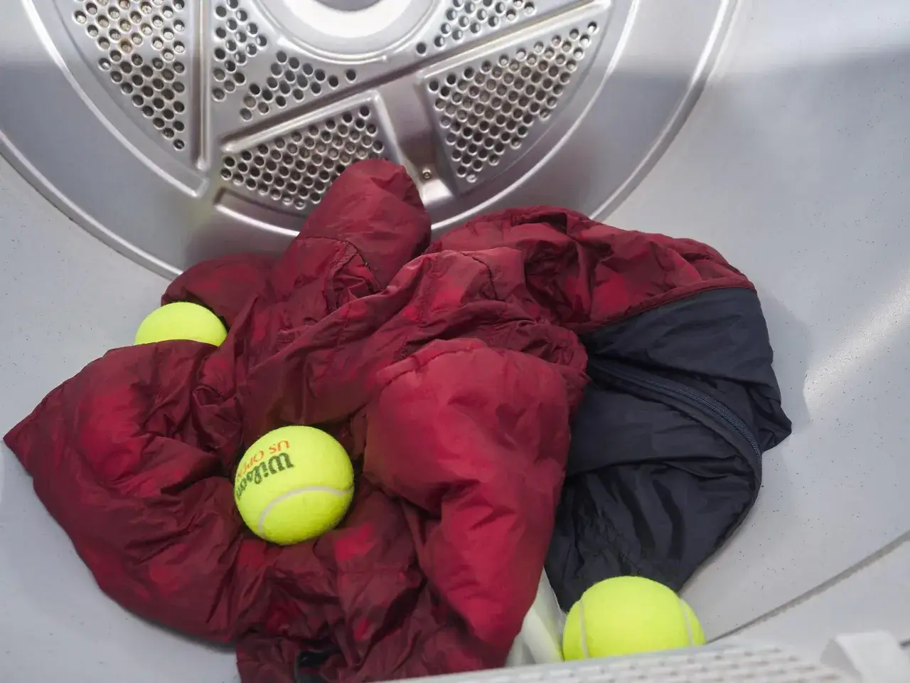 Dentro de una secadora, un edredón rojo y un abrigo oscuro junto a pelotas de tenis. Un truco para como secar un plumas y que quede esponjoso.