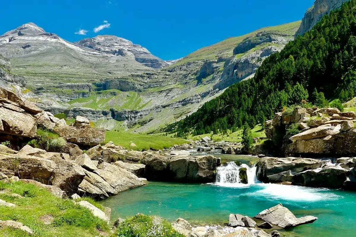 Río turquesa y cascadas en el Parque Nacional de los Pirineos, rodeado de montañas escarpadas y bosques frondosos.
