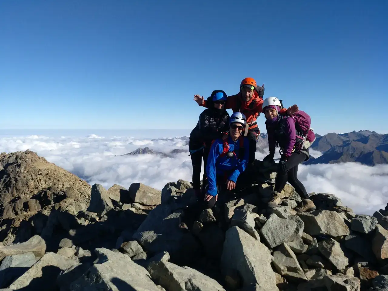 Cinco montañeros celebran en la cima rocosa del Pirineo Occidental, con un mar de nubes bajo un cielo azul.