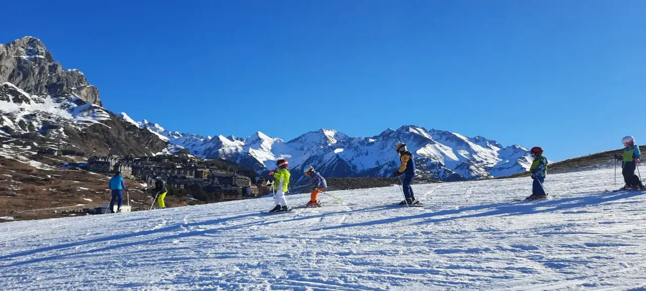 Ni&ntilde;os aprendiendo que es el esqu&iacute; alpino en una ladera nevada, con majestuosas monta&ntilde;as y un pueblo al fondo.