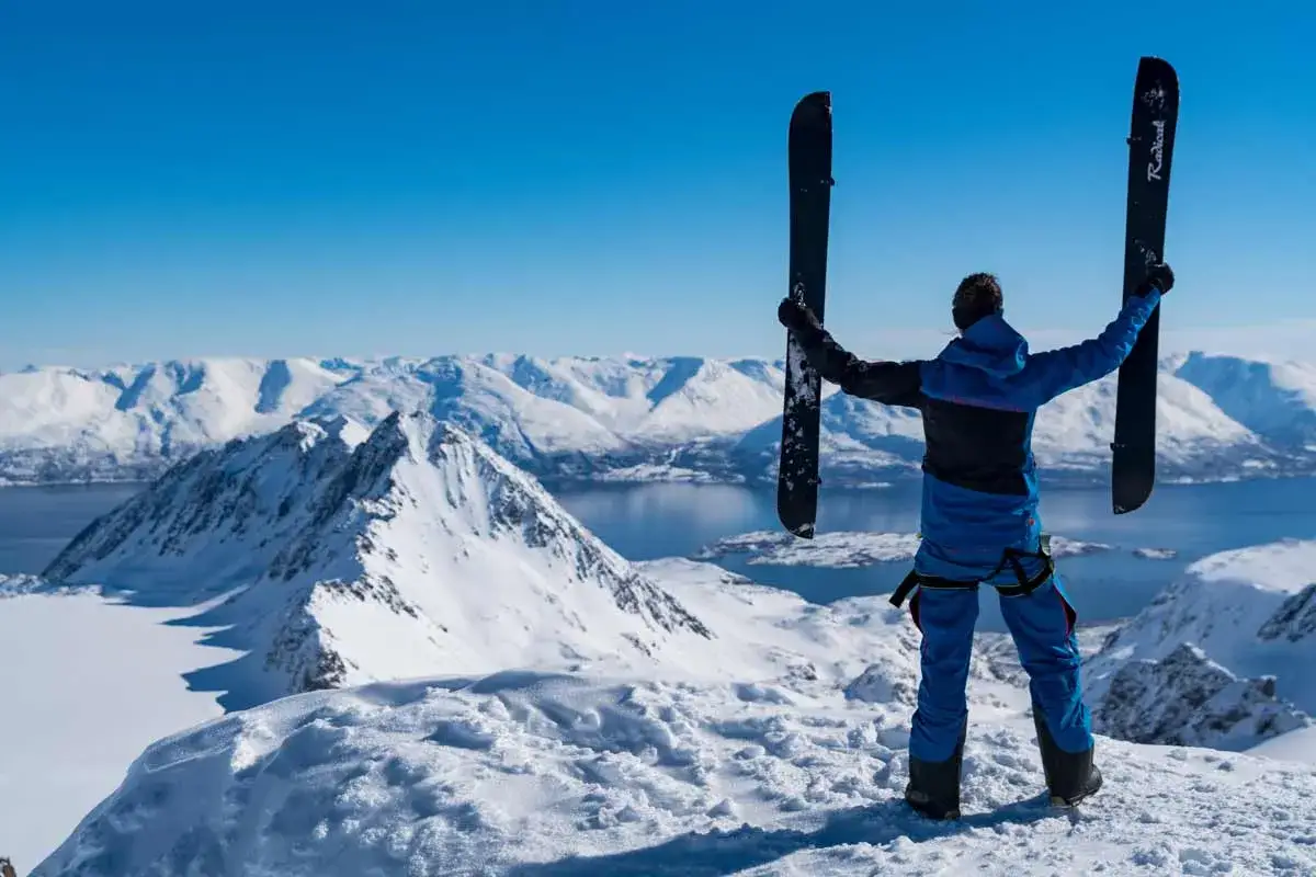 Persona en la cima de una montaña nevada, sosteniendo un splitboard que es, listo para descender. Paisaje alpino espectacular.