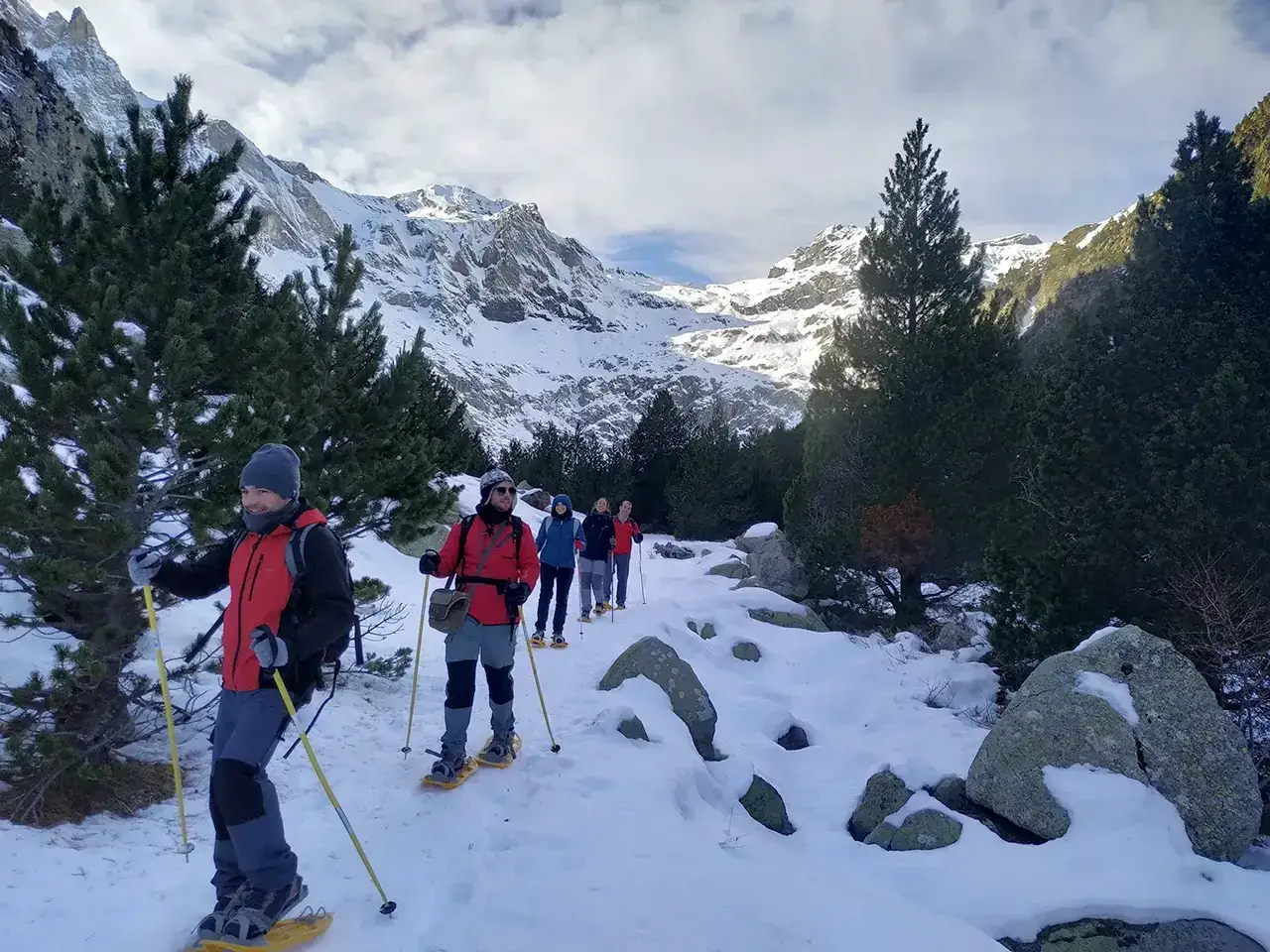 Grupo de personas disfrutando de rutas raquetas de nieve en Cataluña, caminando por un sendero nevado entre pinos y montañas.