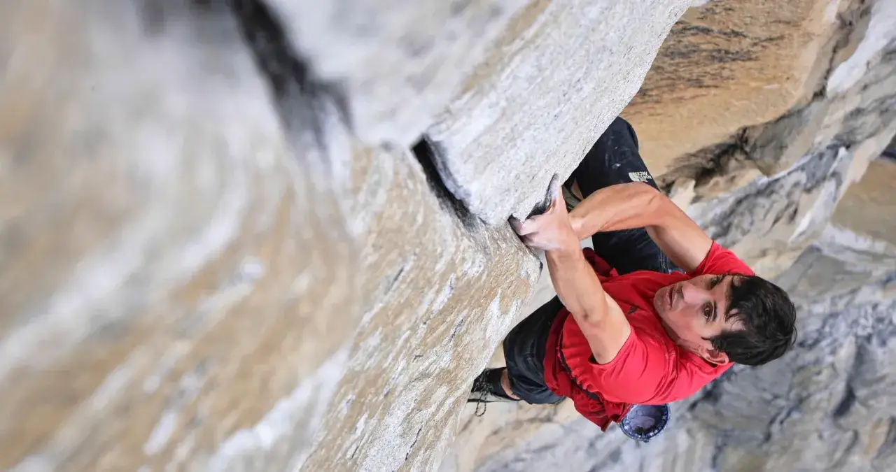 Un escalador con camiseta roja se aferra a una pared de roca, capturado en un momento de tensi&oacute;n para un documental de escalada.