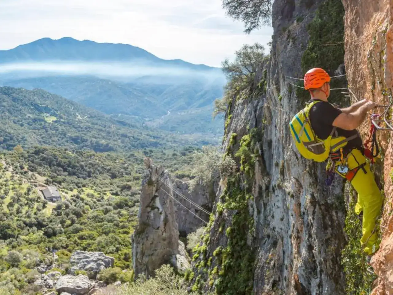 Hombre en equipo de seguridad asciende por una pared rocosa, disfrutando de un paisaje monta&ntilde;oso. Esto es una v&iacute;a ferrata.