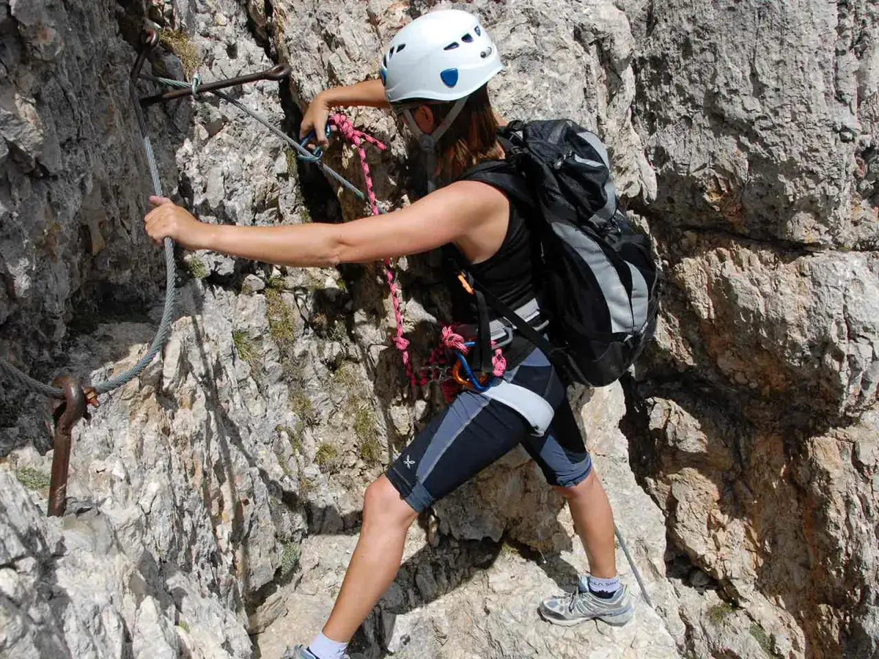 Mujer con casco y mochila asciende por una pared rocosa usando cables de seguridad, disfrutando de los grados escalada.
