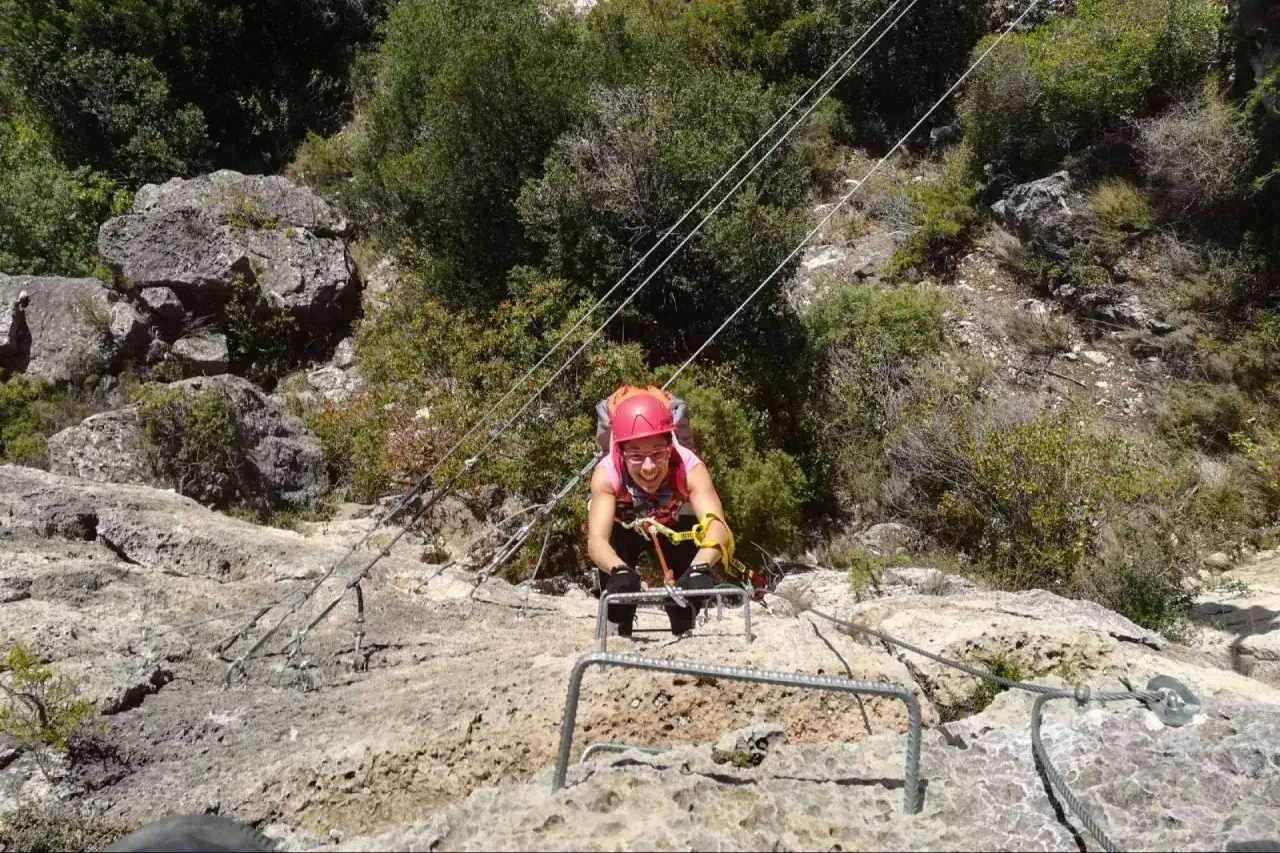 Aventura en una vía ferrata de Catalunya. Mujer con casco y arnés avanza por la roca, asegurada por cables.