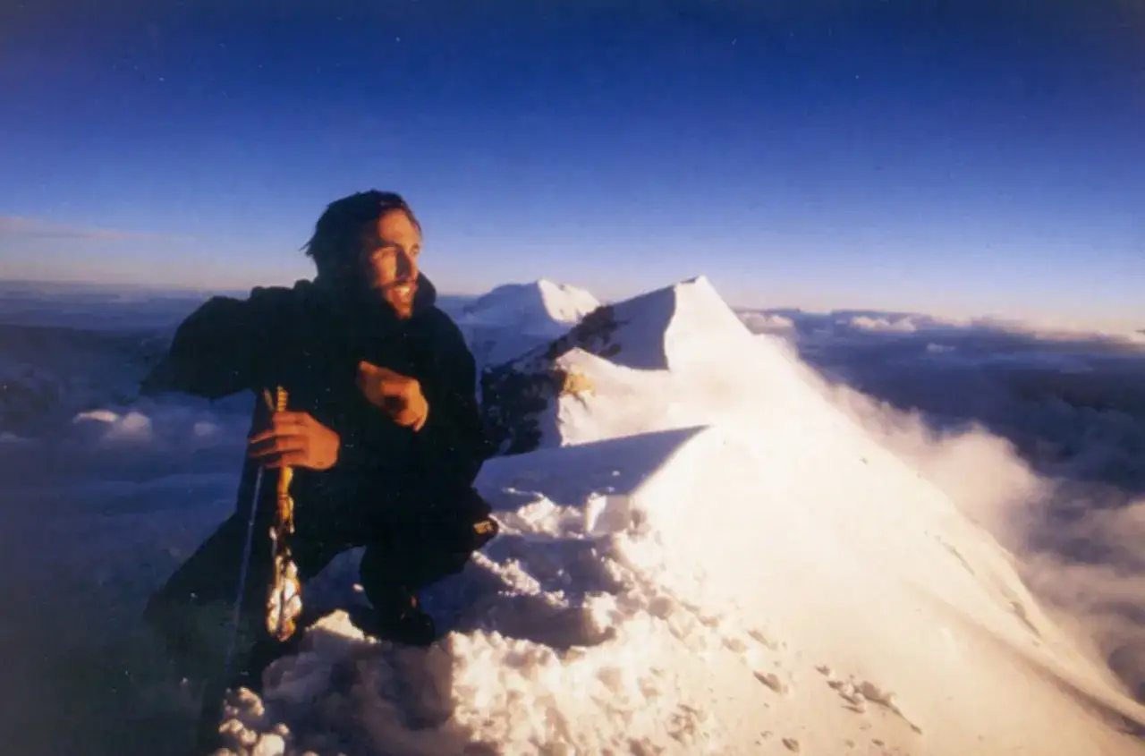 Erik Weihenmayer, sonriente en la cima nevada, con picos y nubes de fondo.