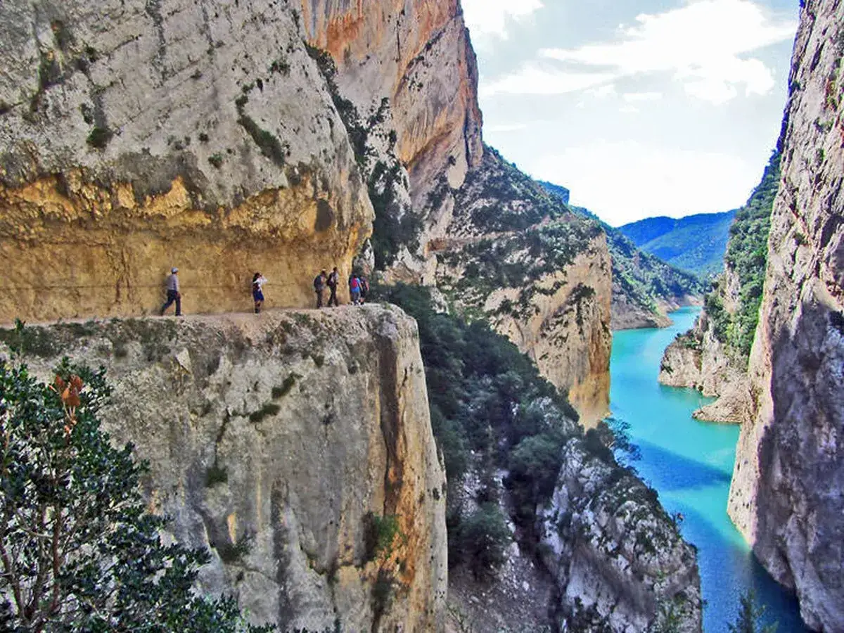 Senderistas caminan por un sendero estrecho al borde de un cañón con un río turquesa. ¡Qué montes bonitos!
