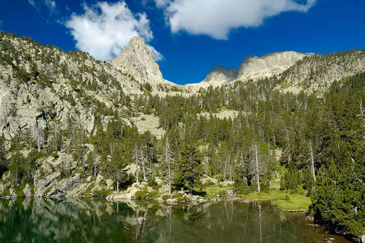 Reflejos en el ibonet de Batisielles, rodeado de montañas rocosas y un denso bosque de pinos bajo un cielo azul con nubes.