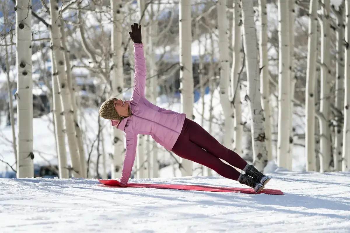 Mujer en postura de plancha lateral sobre una esterilla roja en la nieve, rodeada de árboles blancos. Perfectos ejercicios para esquiar.