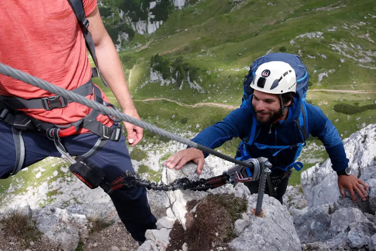 Dos escaladores disfrutan de las vies ferrades en un paisaje montañoso. Uno, con casco y mochila, se agarra a un cable de acero.