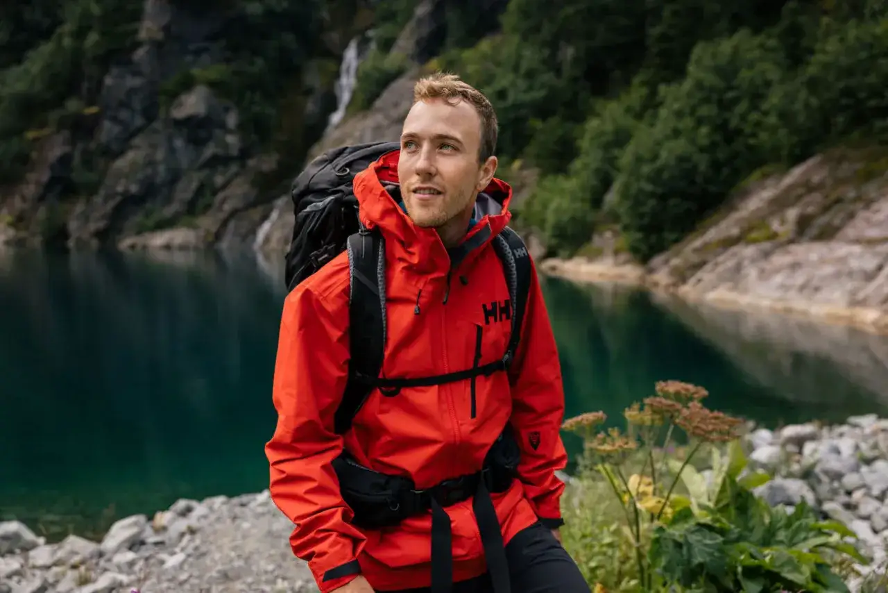 Hombre con chaqueta roja Helly Hansen y mochila, explorando un paisaje montañoso con un lago azul y cascada. La helly hansen historia se vive en cada aventura.