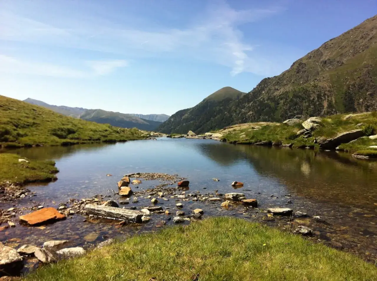 Tranquilo lago de montaña en grp andorra, rodeado de verdes laderas y picos rocosos bajo un cielo azul.