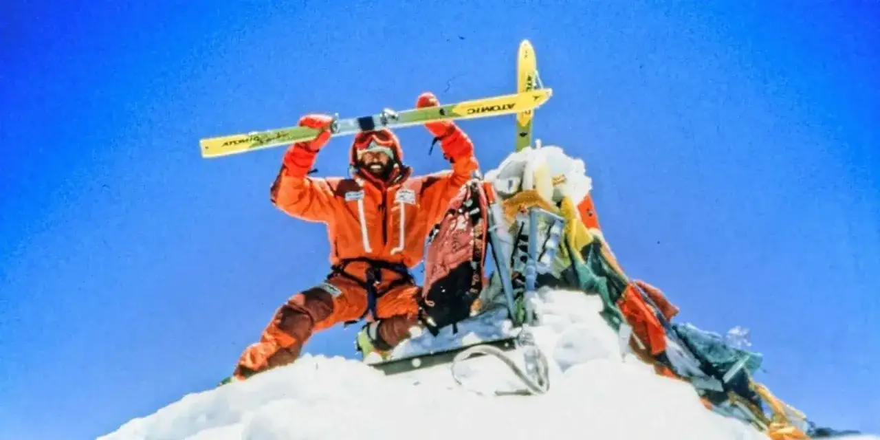 Hans Kammerlander, con esquís, celebra en la cima nevada, con banderas de oración ondeando al viento.