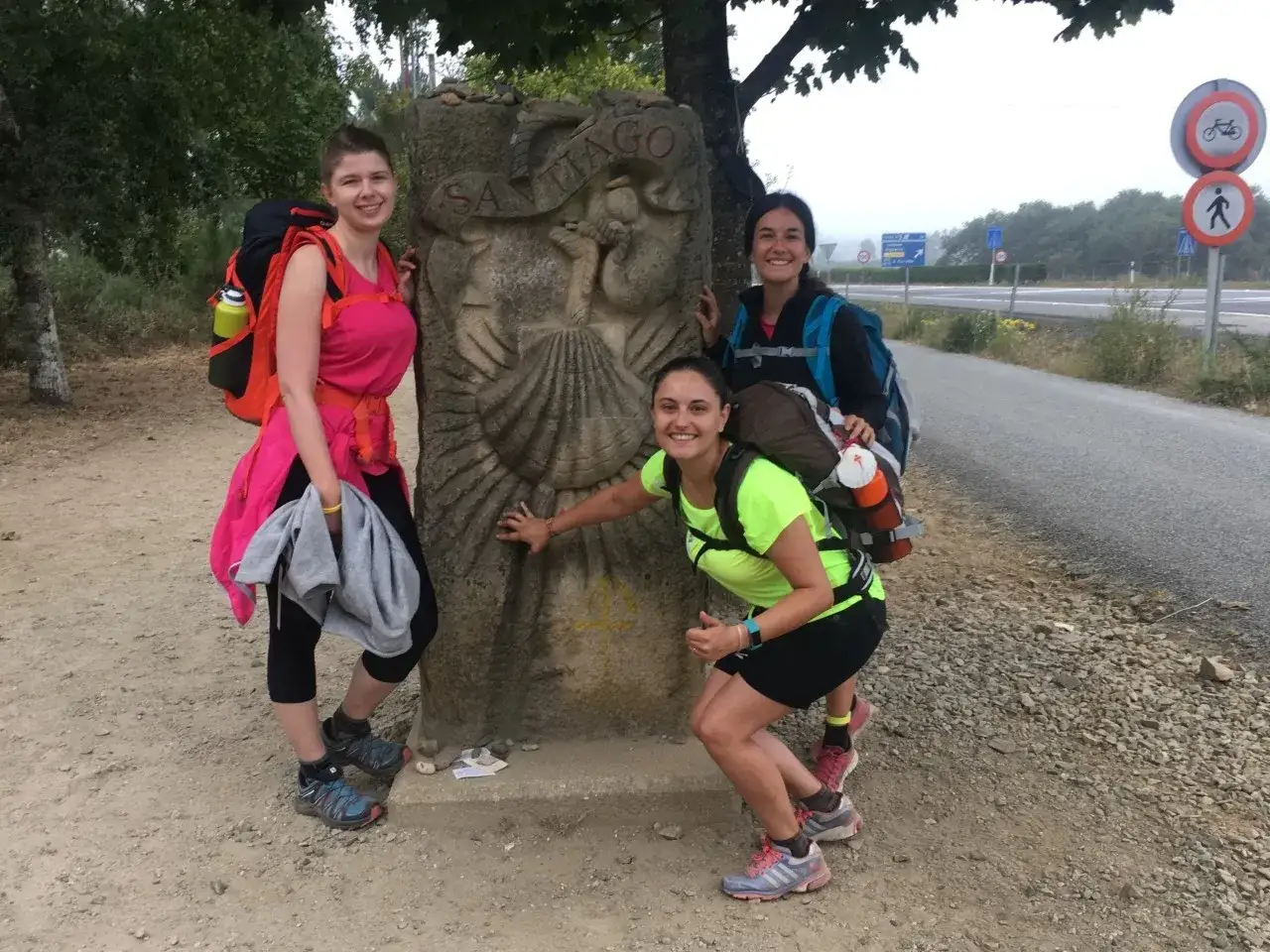 Tres peregrinas sonríen junto a un mojón del Camino de Santiago. El tamaño de su mochila es perfecto para el camino.
