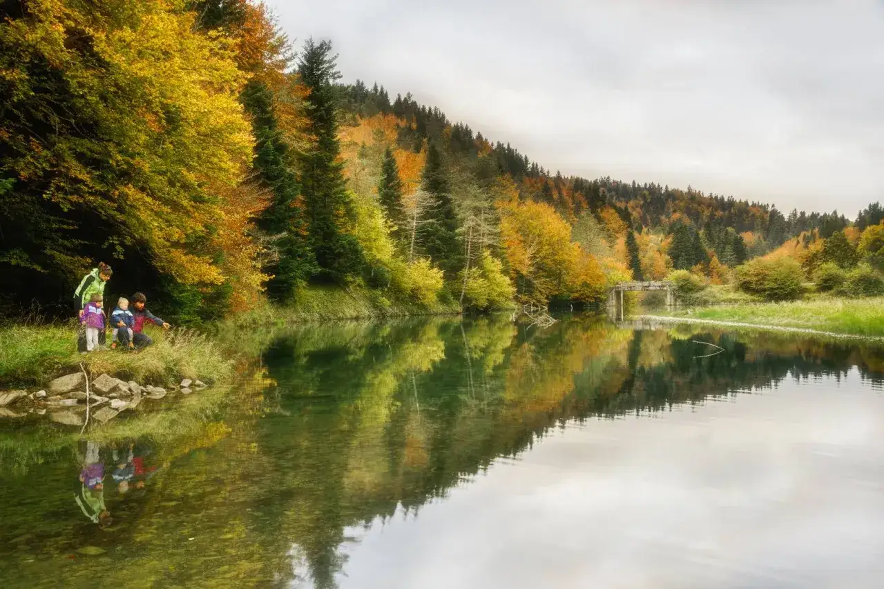 Familia explorando la selva de Irati en otoño, con árboles de colores vibrantes reflejados en un río sereno.