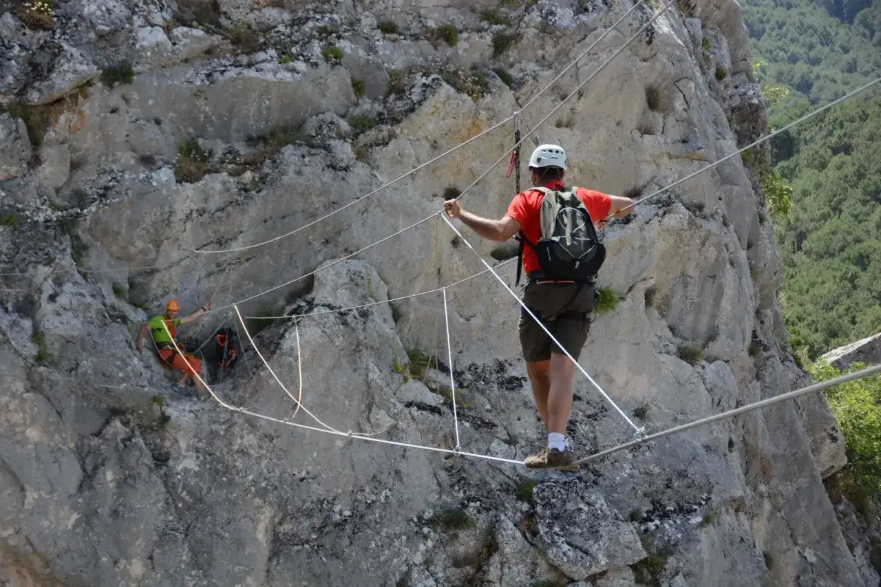 Hombre con casco y mochila cruza un puente colgante, parte del material de v&iacute;a ferrata, sobre un barranco rocoso.