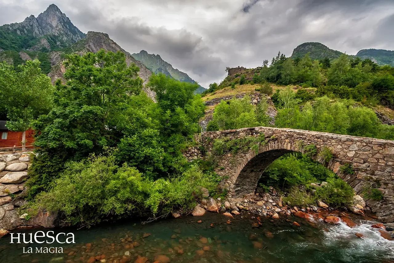 Puente de piedra sobre un río en el valle de Vallibierna, rodeado de exuberante vegetación y montañas imponentes bajo un cielo nublado.