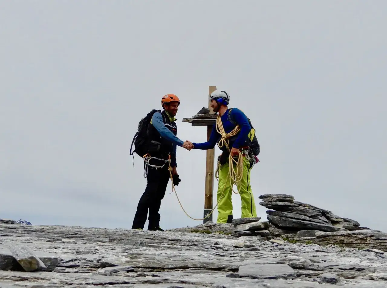 Dos alpinistas se dan la mano en la cima, celebrando su senderismo nieve. Llevan equipo de escalada y mochilas.