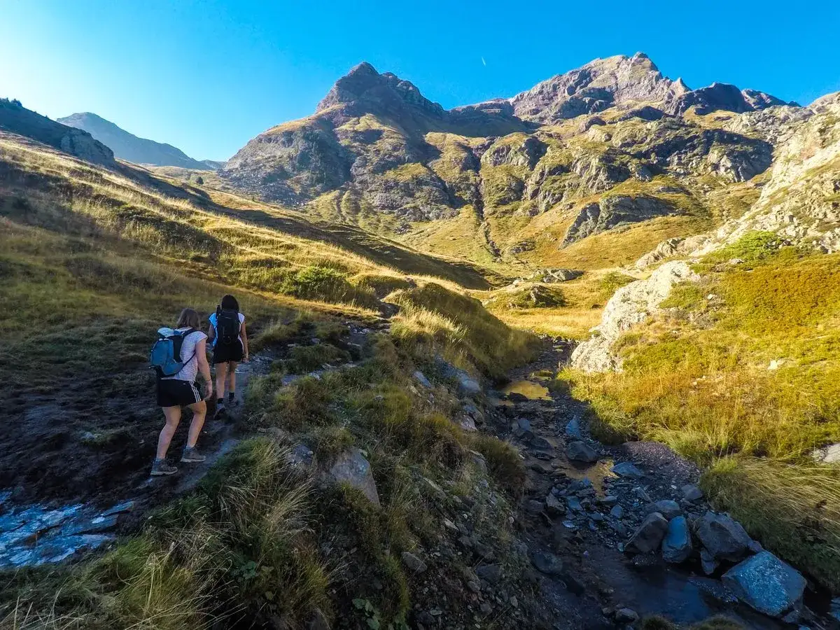 Dos excursionistas suben por un sendero rocoso junto a un canal roya, con imponentes montañas al fondo bajo un cielo azul.