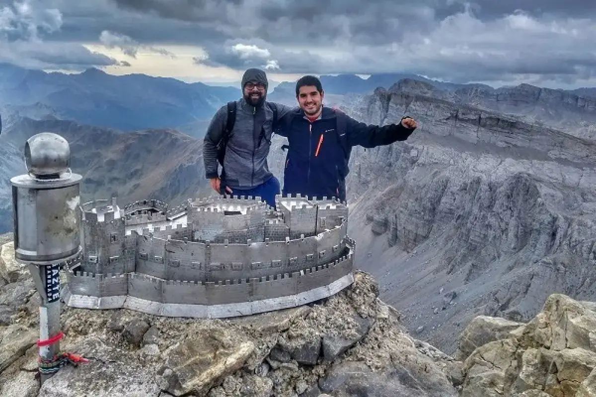 Dos amigos posan junto a una maqueta de un castillo en la cima de una montaña, con un paisaje montañoso espectacular de fondo. El erregeen mahaia parece un tesoro en este lugar remoto.