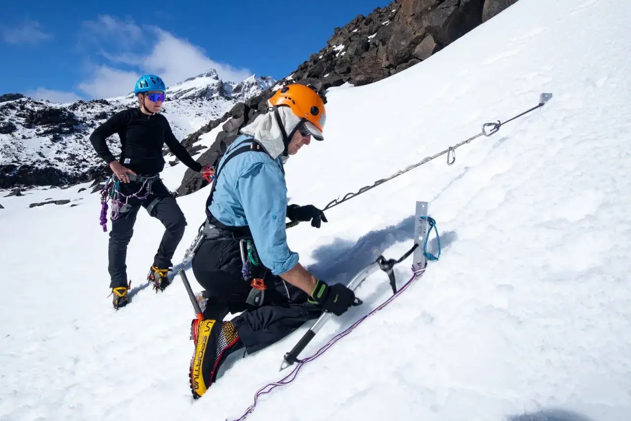 Dos alpinistas se aseguran con una estaca de nieve en una ladera nevada, listos para ascender.