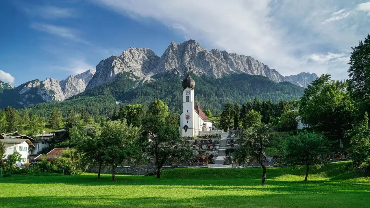 Iglesia blanca con campanario y cementerio en los Alpes alemanes, rodeada de bosques y montañas imponentes.