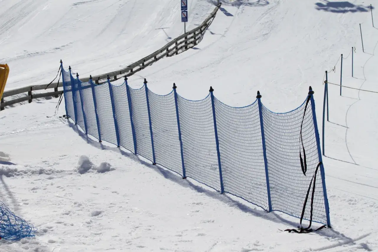 Una valla azul protege la pista de esquí alpino. La nieve cubre la ladera, con huellas de esquí visibles.