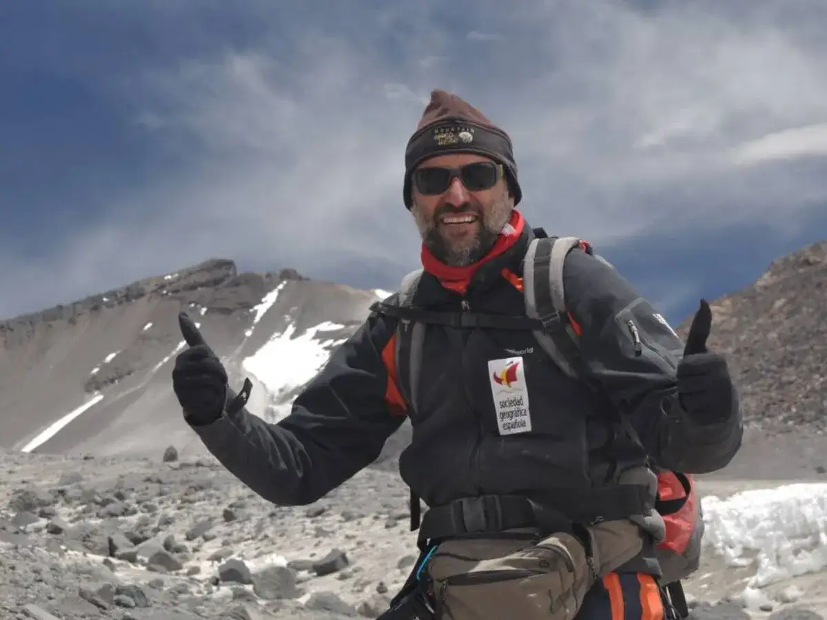 Joao Garcia, sonriente y con pulgares arriba, en la cima de una montaña rocosa y nevada.