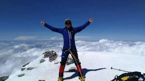 Javier García Díaz, alpinista, celebra en la cima nevada, rodeado de nubes y un cielo azul intenso.